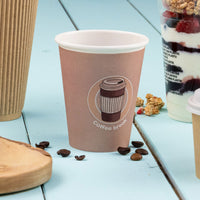 Pappbecher mit rosabraunem Aufdruck und weißem Rand zwischen Kaffeebohnen auf hellblauem Holztisch
